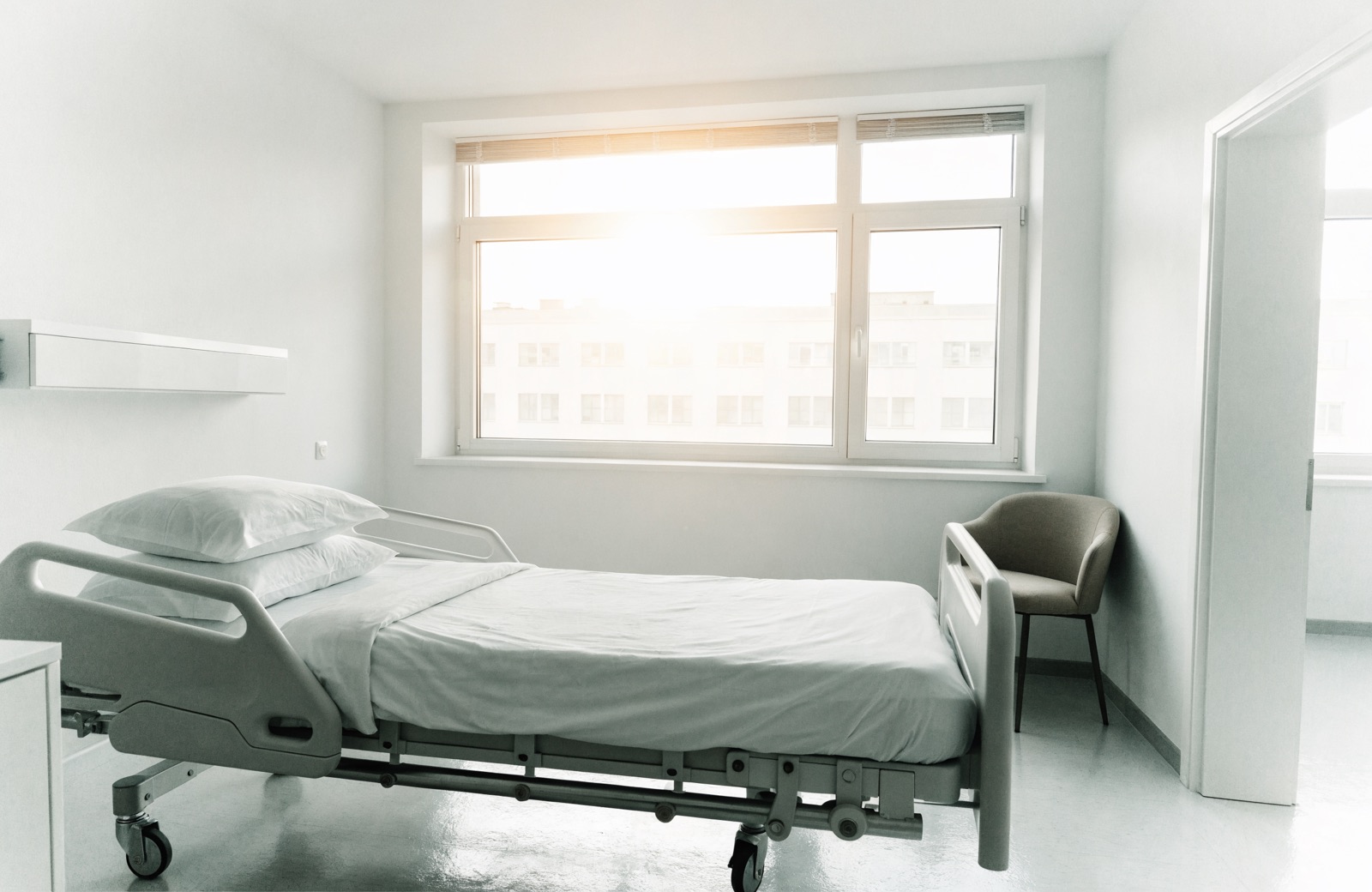 Leeres, frisch bezogenes Krankenbett neben einem Sessel am großen Fenster im Morgenlicht