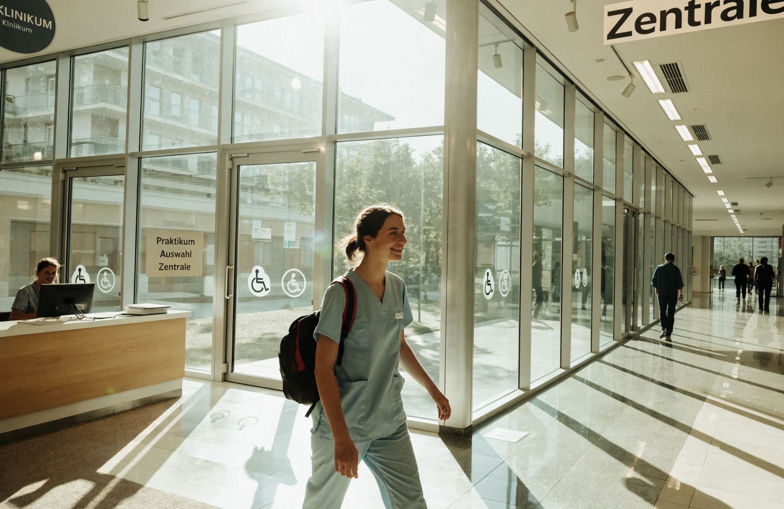 Junge Pflegekraft mit Rucksack geht durch ein helles, verglastes Klinikfoyer